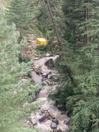 The image depicts a fast-flowing, muddy river winding through a dense forest in Khakhnal, India. The riverbed is rocky, with visible boulders and debris. On the left bank, a bright yellow tarp is partially visible, covering what appears to be a structure or supplies. Tall coniferous trees with dense green foliage line both sides of the river, creating a shaded and somewhat enclosed environment. The water's movement suggests a steep gradient. No people are visible in the scene. The overall impression is one of a natural, rugged landscape with some human intervention indicated by the yellow tarp.