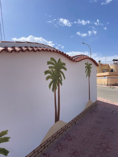 The image displays a bright, daytime scene primarily featuring a white wall adorned with painted palm trees, set against a vivid blue sky. The wall, which appears to curve gently, is topped with distinctive terracotta-colored, wavy roof tiles. Several realistic palm tree illustrations, complete with green fronds and brown trunks, are painted onto the white surface. Two full palm trees are prominently visible, while the fronds of a third can be seen at the bottom of the frame.
Behind the wall, a patterned sidewalk or path is visible, paved with reddish-brown tiles arranged in a hexagonal design. Beyond this, a narrow asphalt road can be glimpsed, leading to a light-colored building in the background. This building, possibly stucco, features a small balcony with a white railing and a prominent band of blue and white decorative tiles along its facade.
Above the architectural elements, a clear blue sky extends across the majority of the upper frame, dotted with scattered, wispy white clouds. A curved street lamp pole reaches into the sky from the background. In the lower-left corner, a few thin black utility wires are also visible. The scene is brightly illuminated by natural sunlight, indicating it is daytime. There are no people or discernible activities taking place, and no visible text. The overall impression is a quiet, sunny streetscape.