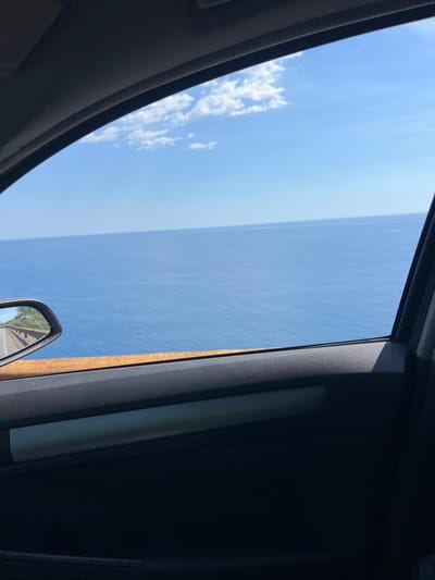 The image captures a view from inside a vehicle looking out of a partially opened window towards a vast expanse of the Adriatic Sea on a clear, sunny day. The water is a deep blue, meeting a pale blue sky dotted with sparse white clouds. In the side mirror, a road and a rust-colored guardrail are visible, suggesting the vehicle is traveling along a coastal route. The interior of the car, including the door panel and a reflective strip, is also visible in the foreground, framing the scenic ocean view. The location is identified as Dobra Voda, Montenegro.