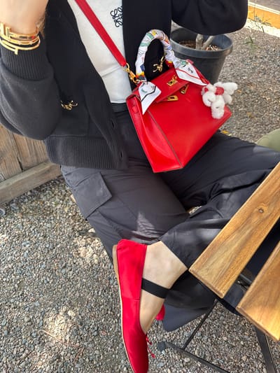 The image shows a person seated at an outdoor table on a gravel-covered surface. The person is wearing dark pants and red ballet flats with black straps. On their lap sits a red handbag with gold hardware, adorned with a white scarf and a small, white, fuzzy decoration. Part of a black and white top or sweater is visible, featuring a Loewe logo. A gold bracelet is visible on the person's wrist. A wooden table partially obscures the view of the person's legs. There is a plant pot in the background. The image suggests an outdoor setting, possibly a patio or garden. The specific location within the city of Chula Vista is not discernible from the image.
