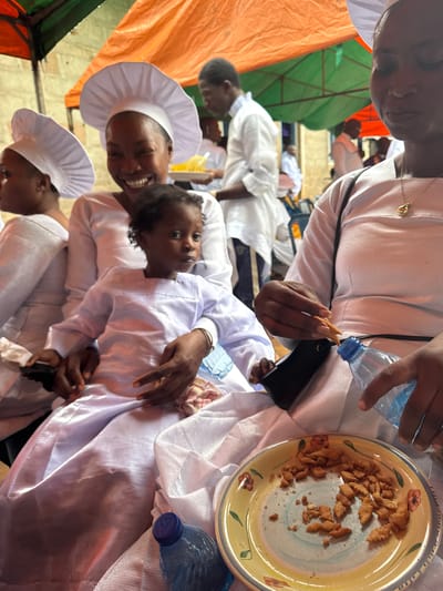 In Ikoga, Nigeria, a group of people, some wearing white priestly attire and head coverings, are gathered under a colorful awning. A woman with a bright smile holds a young child, also dressed in white. In the foreground, a plate of small, baked snacks sits next to a bottle of water. The woman's hand is reaching for a snack, and another person's hand is visible, holding a piece of food near the water bottle. The overall atmosphere appears to be one of communal gathering or celebration.