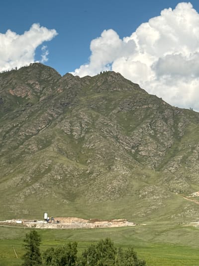 The image depicts a vast, grassy mountain range under a partly cloudy blue sky. The mountains are covered in green vegetation and rocky outcrops. In the foreground, a green field leads up to a construction site at the base of the mountains. The construction site features several white silos, a yellow excavator, and piles of dirt and building materials. A few trees are visible in the very bottom of the frame. The scene appears to be located in a rural or semi-rural area, possibly on the outskirts of the city of Onguday, Russia, given the context provided. There are no people visible in the image.