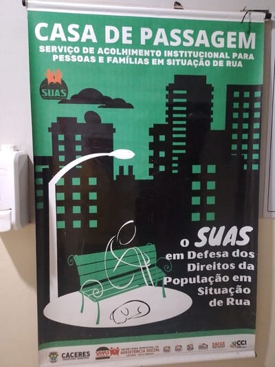 The image shows a poster displayed on a wall, likely in Cáceres, Brazil. The poster is primarily green and black and features text and graphics related to a "CASA DE PASSAGEM" (Shelter House) which offers institutional support for people and families on the street.

The poster has a large white title at the top: "CASA DE PASSAGEM SERVIÇO DE ACOLHIMENTO INSTITUCIONAL PARA PESSOAS E FAMÍLIAS EM SITUAÇÃO DE RUA". Below this, a graphic depicts a stylized cityscape with silhouetted buildings. In the foreground, a white street lamp curves over a green park bench where a white line drawing of a person is seated. A small outline of a dog is drawn near the bench. To the right of the bench, white text reads: "o SUAS em Defesa dos Direitos da População em Situação de Rua".

At the bottom of the poster, there are several logos and names, including "CACERES PREFEITURA MUNICIPAL", "SMAS", and other organizational identifiers. The context suggests this is a public service announcement or information poster within the municipality of Cáceres.