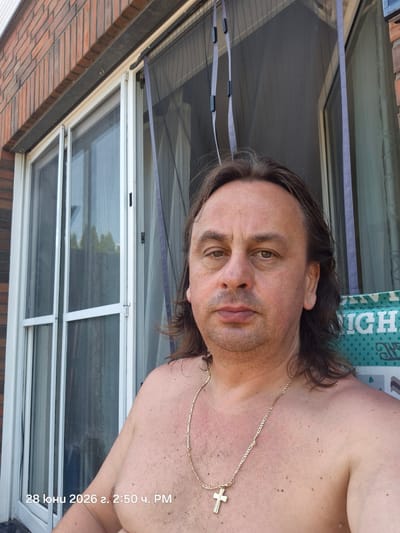 A man with long, dark hair, shirtless, is shown from the chest up, with his head tilted to his left. He is wearing a thin gold chain with a cross pendant. His skin has a reddish hue, possibly from sun exposure, and there are scattered moles on his chest.
The man is outdoors, with a window and screen visible behind him, suggesting a residential setting. To his left, a brick wall is visible. Above him, a part of a tiled roof can be seen. The lighting suggests it is daytime.
The image has text overlaid on the right side, which appears to be a date and time stamp in a non-English script: "28 юни 2026 г. 2:50 ч. PM". This indicates the photograph was taken on June 28, 2026, at 2:50 PM. The overall impression is a casual, personal snapshot taken outdoors during daylight hours.