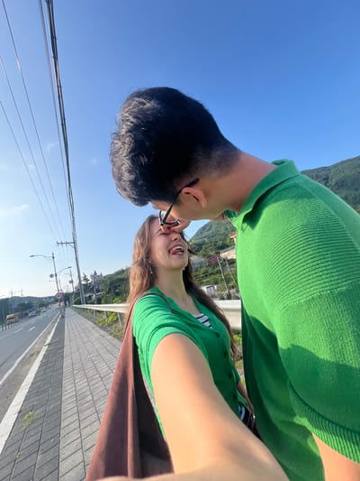 This is a vertical, selfie-style photograph featuring two young people, appearing to be a couple, in a close embrace. The man, on the left, has short, dark hair and is wearing a bright green sweater. The woman, on the right, has long, light brown hair and is wearing a green top with black and white stripes visible underneath. She is wearing glasses and her mouth is open in a happy expression, suggesting laughter or joy. They are positioned so their faces are very close, with the man leaning in towards the woman.
The background shows a clear, bright blue sky, indicating a sunny day. Below them, a landscape unfolds with green hills dotted with buildings, suggesting an elevated viewpoint overlooking a town or village. A paved pathway with distinct grey paving stones runs alongside a road, where a few cars are visible. Power lines stretch across the sky, supported by poles. A circular speed limit sign with "40" is visible on a pole, along with streetlights. The overall scene suggests an outdoor, possibly scenic location. The time of day appears to be daytime, given the bright sunlight and clear sky. The mood conveyed by the couple's interaction is affectionate and joyful. The image is taken from a high angle, looking down at the road.