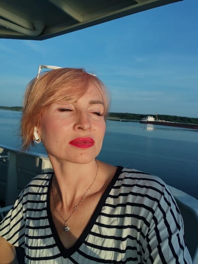 A woman with short, blonde hair and bright red lipstick is taking a selfie on a boat. She is wearing a black and white striped shirt and a silver necklace with a crown pendant. Her eyes are closed, and she is looking towards the right side of the frame. In the background, there is a wide body of water with a barge in the distance and a treeline on the horizon. The sky is a clear blue. The boat appears to be on a river or canal in Shchetinino, Russia.