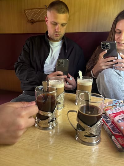 In a train cabin, two individuals are seated at a wooden table. One person is holding a smartphone, while the other is stirring a drink in a glass with a metal holder. A second glass with a metal holder sits beside it. A packaged food item lies on the table. The seat is upholstered in a dark red material, with a rack on the wall. Based on the location, this is likely a scene from a train journey through Тарноўскі сельскі Савет, Belarus.