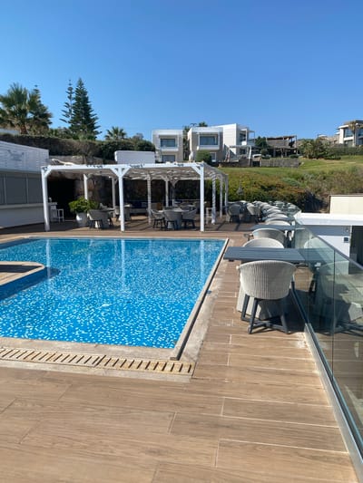 The image depicts an outdoor seating area next to a swimming pool. The swimming pool is tiled with blue and white mosaic tiles and has a wooden deck surrounding it. There are several tables and chairs set up under a white pergola, with some tables also along a glass railing overlooking the pool. In the background, there are modern white buildings and lush green landscaping, with a clear blue sky above. The scene appears to be at a resort or hotel in Panormos, Greece, given the architectural style and vegetation typical of the region. There are no people visible in the image.