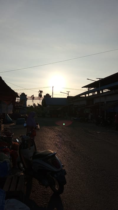 The image depicts a street scene at sunset in Puloara, Indonesia. The sun is low in the sky, casting long shadows and a warm glow over the asphalt road. On the left side of the road, there are market stalls with awnings and various goods, possibly produce, piled on wooden pallets. A scooter is parked in the foreground, its license plate visible.
In the middle ground, a person wearing a headscarf is walking away from the camera, carrying a plastic bag. Beyond them, there are buildings that appear to be part of a market or commercial area, with some palm trees and a decorative archway visible in the distance. The sky is clear with a gradient of pale blue to yellow near the horizon, indicating the setting sun. The overall atmosphere is quiet and tranquil, characteristic of the end of the day in a local Indonesian community.