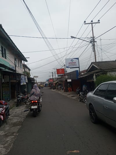 This image depicts a street scene in the city of Houaythat, Laos, under an overcast sky. The view is from a low angle, looking down a paved, two-lane road flanked by buildings and utility infrastructure.
In the immediate foreground on the left, a red scooter is partially visible on the sidewalk, with a portion of its license plate "D 43 YG" legible. Adjacent to it, another dark-colored motorcycle is parked on the grey concrete sidewalk. On the same side, a two-story building features several signs, including "Mini ATM" and services such as "TRANSFER TARIK TUNAI PULSA KUOTA PRINT A3 CETAK FOTO SCAN LAMINATING," along with "BDS" and "OVO" logos.
To the right of the center, a person is riding a white scooter away from the viewer. The rider wears a patterned head covering (hijab) and a patterned lower garment, possibly a long skirt or sarong over pants. The scooter's license plate reads "F 6872 HBN" with "09 30" below it, and its rear brake light is illuminated. Further right, a silver sedan is parked parallel to the curb, displaying black multi-spoke rims on its visible rear wheel.
The midground features a dense array of commercial and residential buildings on both sides of the street. On the right, a prominent white sign with blue lettering identifies "APOTEK PRONESIA" and a medical cross logo, with smaller red text underneath. Next to it is a smaller red sign for "TANIA." A utility pole with multiple cross arms supports a street light and a dense tangle of overhead electrical and communication wires that crisscross the sky. A faint communication tower is visible in the distant background. On the left midground, additional shops and buildings are present, with visible signs including "PERUM" and "BCA," and one noting "PHOTO COPY." Several other motorcycles are parked along the sidewalks.
The street surface is asphalt, showing some wear and minor debris. The general environment suggests a bustling urban or semi-urban commercial area.
