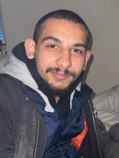A single male individual is depicted in a frontal close-up, situated indoors in Brežice, Slovenia. The subject exhibits features commonly associated with Middle Eastern or South Asian ethnicity, possessing dark brown eyes, short dark hair, and a trimmed dark beard and mustache. A small pigmented lesion is present on the left upper lip. He maintains direct eye contact with the camera and displays a slight smile. The individual is wearing layered garments: a dark (possibly black or very dark grey) outer jacket, a navy blue puffer vest with a visible orange interior lining, and a black hooded sweatshirt with a light grey hood and white drawstrings. The background consists of a plain, light-colored wall. A vertical window blind is partially visible in the upper left corner, and a crumpled white textile is visible on the right side of the frame. Illumination appears to be artificial.