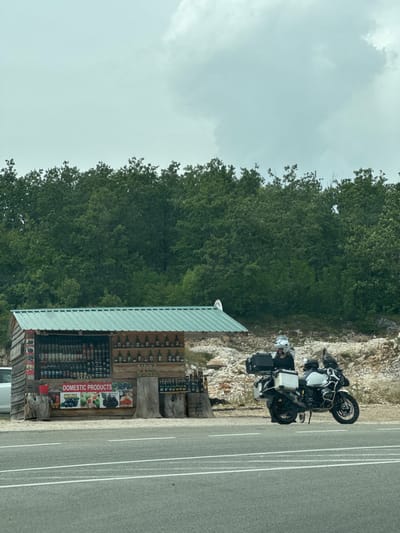 The image shows a roadside stall selling "DOMESTIC PRODUCTS" with a motorcycle and a person parked next to it. The stall is made of wood and has a green metal roof. It is stocked with numerous bottles and jars of various sizes and colors, appearing to be preserves, liqueurs, or juices. A banner on the stall displays images of fruits like plums, figs, and berries, suggesting these are some of the products offered. To the right of the stall, a large, white motorcycle with saddlebags is parked. A person wearing a helmet and motorcycle gear is standing next to the motorcycle, appearing to be engaged with it. The background consists of a dense forest of green trees, with a rocky, uneven terrain behind the stall. The scene is set on the side of a paved road with white lane markings. The sky is overcast with some clouds. This location is identified as Jasenovo Polje, Montenegro.