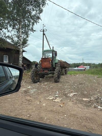 The image depicts a rural scene in the city of Yug, Russia. A green tractor with a rusted front and a trailer is parked on a dirt road. The trailer is partially filled with wood. To the left of the tractor is a wooden house with a dark roof and a visible window. Two adults and a child stand near the house, looking towards the tractor. A car's side mirror is visible in the foreground on the left, reflecting a portion of the scene, including the house and the sky. Power lines and poles are prominent in the background, with a line of trees and a few scattered houses visible further in the distance. The sky is overcast.