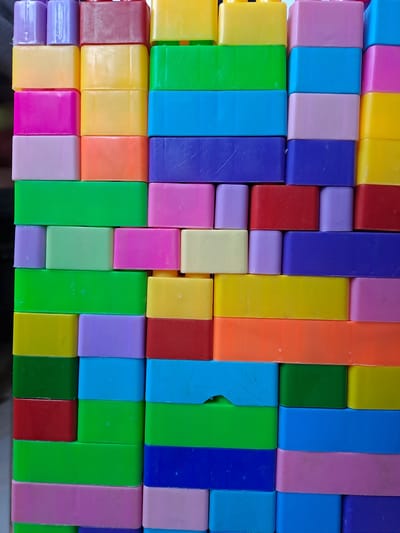 This is a close-up, eye-level, full-frame shot of a collection of colorful, rectangular building blocks arranged in rows. The blocks are made of plastic and come in a variety of vibrant colors, including red, green, yellow, blue, purple, pink, and orange. The blocks are arranged in a staggered pattern, with some rows shifted slightly to the left or right relative to the rows below. The lighting appears to be natural and even, suggesting the image was taken in a well-lit indoor environment. There are no people or animals visible in the image, and the background is blurred, focusing all attention on the blocks themselves. The arrangement of the blocks creates a sense of order and playfulness. The surface on which the blocks are placed is not visible. The context provided suggests this image was taken in Lhokaweawe, Indonesia.