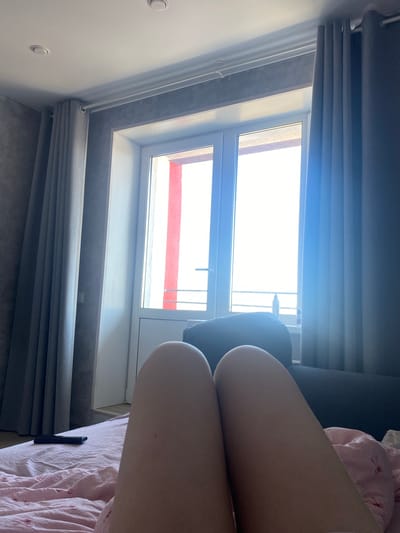 The image captures an indoor scene, likely a bedroom or living room, from a low perspective, rotated 90 degrees counter-clockwise. In the foreground, a person's bare legs, specifically the thighs and knees, are visible, resting on pink and white patterned bedding. The legs extend from the right side of the frame.
A large, modern window with white frames dominates the left and central portion of the background. The view outside the window is extremely bright, indicating strong daylight. Below the window, gray pleated Roman blinds are visible, partially drawn. Similar gray curtains are present above the window, also partially covering the top part of the frame. A dark piece of furniture, possibly a sofa armrest or headboard, is partially seen behind the legs, near the right side of the window. On the white wall to the far left, two recessed circular light fixtures are visible. Below the window, a windowsill and a darker, textured wall or floor material are present. A small black rectangular object, likely a phone or remote control, rests on the pink bedding near the bottom right.
The activity depicted is a person resting or relaxing indoors during the daytime in Zubovo, Russia. No visible text is present.