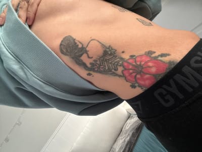A close-up indoor shot shows a person's torso, specifically the side and hip area. A black waistband with the word "GYM" visible sits atop the hip. A large tattoo is present on the skin, featuring a red flower and a darker, abstract design. The person is wearing a light blue or grey sweatshirt. The background is out of focus, but hints of a pillow and what seems like a white tiled wall are visible. The location of the image is Seal Beach, United States of America.