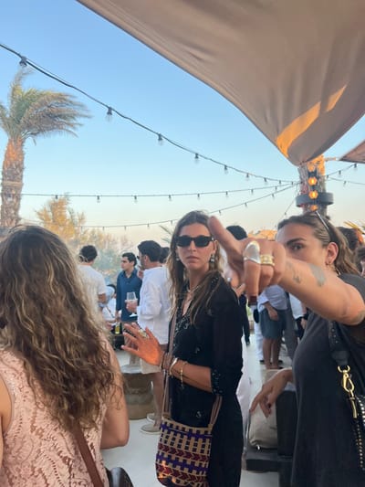 The scene depicts an outdoor social gathering at a venue in Psarou, Greece, during late afternoon or early evening under a clear blue sky. Overhead, strands of festoon lights with exposed bulbs are strung across the area. A large, light-colored fabric canopy extends from the upper right, partially shading the space. To the far left, a tall palm tree is visible.
In the immediate foreground, the back of a person with long, wavy brown hair, wearing a light pink or peach lace top, is positioned on the left. To their right, a woman with long dark hair and rectangular sunglasses stands facing slightly left, looking directly towards the camera. She wears a black long-sleeved top with subtle embellishments, numerous rings on both hands, multiple bracelets on her left wrist, and a patterned woven cross-body bag with tan, purple, and black tones. Her right hand is slightly raised.
To the right, a second woman is partially visible, facing slightly left with her head turned towards the camera. She has light brown hair, small hoop earrings, and wears a dark top. Her right arm is extended forward, with the index finger pointing directly towards the viewer. Multiple rings are visible on her fingers, including a prominent silver one and several gold-toned bands. A tattoo is discernible on her right forearm or hand.
In the mid-ground and background, several individuals, predominantly men in light-colored shirts (white, light blue), are observed socializing. One person in the background holds a clear glass containing a drink, possibly wine. A wooden post with circular orange lights is situated on the right side of the frame, behind the pointing woman. The environment indicates a relaxed, open-air setting.