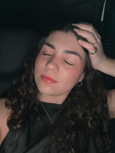 A young woman lies with her eyes closed. She wears dark clothing and has long, curly dark hair. Her left hand rests on her head, adorned with a few rings. Her skin appears smooth, with a touch of blush on her cheeks and glossy red lipstick. A delicate silver necklace is visible around her neck. The background is a dark, solid color. The image is softly lit and intimate.