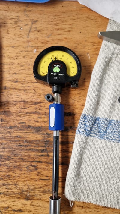 This image shows a close-up, top-down view of a dial indicator gauge and a part of a precision measuring tool. The dial indicator, labeled "Mah. Millimess 1003," has a yellow face with black markings indicating measurements in micrometers (µm), ranging from 0 to 50 in both clockwise and counter-clockwise directions. The needle is currently pointing slightly past the 10 µm mark on the clockwise side. The gauge is mounted on a silver-colored rod which extends to the right. A blue plastic sleeve with a silver insert is attached to the rod. Further along the rod, a section of a metal tube is visible, with the word "DIATEST" imprinted on it. The tools are resting on a wooden surface, likely a workbench, which shows grain and some wear. In the upper part of the image, a piece of textured, white fabric with a blue woven pattern is partially visible, suggesting the presence of other items or materials in the background. The lighting appears to be overhead, casting subtle shadows. The overall scene suggests a workshop or precision measurement environment, possibly in Ichenhausen, Germany, given the location context. There are no people visible, and the focus is solely on the measuring instruments and their immediate surroundings.