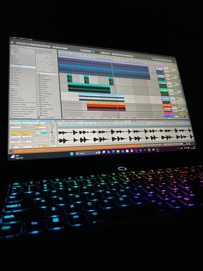 The image depicts a close-up, angled view of an active laptop in a dark environment, presumably in the city of Tairov, Armenia. The laptop screen displays the Ableton Live 12 Suite digital audio workstation (DAW) software, showing multiple tracks with audio waveforms and MIDI clips, indicative of music production activity. The left side of the screen shows the software's browser, while the main window features an arrangement view with several colored tracks, including one highlighted as "025 a. Backing Loop 122bpm Am - LUCIDDEEPMELODIC". The Windows 11 taskbar is visible at the bottom of the screen, displaying a search icon, a weather icon showing "10°C Light rain", a browser icon, and a Spotify icon, along with the date "29.10.2023". Below the screen, the laptop's keyboard is prominently featured, illuminated by a multi-color RGB backlight that transitions from cyan and blue on the left to green, yellow, orange, and purple hues on the right. No individuals or direct human actions are visible, but the active screen and illuminated keyboard suggest recent or ongoing use. The surrounding area is uniformly dark, indicating a low-light setting.