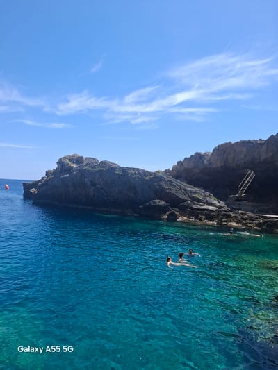 The image captures a coastal scene near Plakias, Greece, with a prominent rocky cliff dominating the center. The cliff face is dark and rugged, showing signs of erosion. A narrow stairway is visible leading up the cliff.

Below the cliff, the turquoise water is clear, revealing varying depths and textures on the sea floor. Three people are swimming in the water.

The sky is a bright blue with thin, wispy clouds streaking across it. A red buoy floats in the water near the bottom of the frame. "Galaxy A55 5G" is written on the right side of the image.