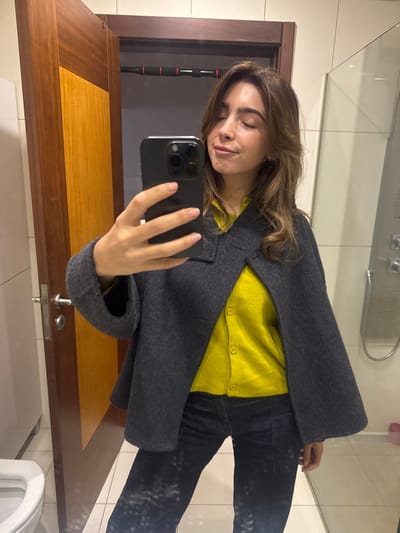 A young adult female, depicted from the mid-thighs up, is taking a mirror selfie in what appears to be a bathroom in Nogueira e Silva Escura, Portugal. She has shoulder-length brown hair, closed eyes, and a slight smile. She wears a bright yellow collared shirt or cardigan, an open dark grey short jacket, and dark blue jeans. Her left hand holds a dark-colored smartphone with a triple-lens camera array, positioned to capture the reflection. The background visible in the mirror shows a white ceramic toilet bowl partially on the lower left. A prominent wooden door with a rectangular yellow panel and a silver handle is visible behind her. Above the door, a black pull-up bar with red accents is mounted in the doorway. To the right, a glass shower partition and a metallic shower control panel are present. The room features light-colored wall tiles and white floor tiles, with a small pink object visible on the shower floor.