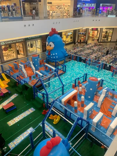 The image displays a multi-level shopping mall environment, specifically an entertainment area located on the ground floor of a mall in Lauro de Freitas, Brazil. A large children's play zone, primarily blue, orange, and green, occupies the central and foreground sections.
**Subjects and Objects:**
The play zone features an extensive inflatable obstacle course with slides, tunnels, and climbing elements in blue and orange. A large ball pit filled with numerous light blue and white plastic balls is integrated into the play area. A colossal inflatable blue chicken character with a red comb and wattle stands prominently at the rear of the play zone, overlooking the ball pit. A partial view of another similar blue inflatable is visible in the foreground. Adjacent to the inflatable structures, a section covered in green artificial turf contains small red and blue tables with benches, patterned ground mats, and three rocking horses (two yellow, one red). The entire play area is enclosed by blue-framed netting barriers.
**Environment:**
The surrounding mall features multiple levels with glass railings. Various retail storefronts are visible on both ground and upper floors, identified by signage including "CAROLINA COSTA," "Ernesto," "Useflex," and "Magazines Luiza." To the right of the play area, a substantial grid of tables displays merchandise, suggesting a temporary book sale or market. The mall is illuminated by overhead lighting fixtures.
**People, Actions, and Interactions:**
Several individuals are observed on the upper mall levels, walking along the walkways. Within the ground-level play area, a limited number of children and adults are present, interacting with the inflatable equipment and within the ball pit. Individuals are also visible near and inside the retail establishments.