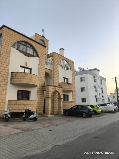 A three-story building with light brown brick accents frames the left side, connected to a taller white building on the right. Several parked cars line the street, including a dark green sedan, a bright green compact car, and other vehicles further down the block. Two scooters sit near the first building. Overhead utility lines crisscross near the taller building, set against a pale sky. The date and time stamp "23/11/2025 06:34" are visible at the bottom right corner of the frame.