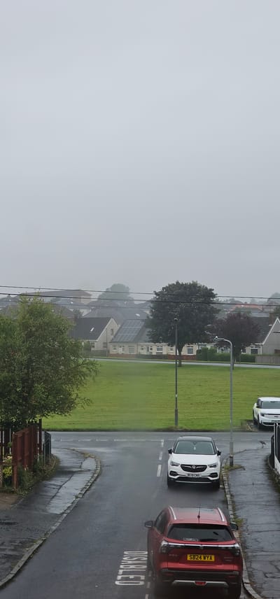 The image depicts a view of a residential street in Kilwinning, United Kingdom, on an overcast day, with visible rain creating wet surfaces. In the foreground, a red Suzuki Vitara SUV is parked on the street, with the word "DISABLED" stenciled in white on the wet asphalt behind it. Further up the street, a white Vauxhall Grandland X is parked. Beyond the street, a large, manicured green space is visible, bordered by houses and trees. Some houses in the background are equipped with solar panels. Power lines stretch across the scene, and a lamppost stands near the green space. A bird is visible on the pavement near the white car. The sky is heavily overcast, obscuring the horizon and lending a muted quality to the scene.