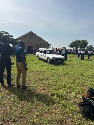 The image depicts an outdoor gathering under bright daylight, likely in a rural or semi-urban setting in Panyam, Nigeria. The scene is dominated by a wide, grassy field with sparse green growth, suggesting dry ground. The sky is clear and blue, indicating sunny weather.
In the foreground, two men stand facing away from the camera. One wears a blue polo shirt and tan trousers, holding a dark object (possibly a phone) in his hand. The other, partially obscured, is in dark clothing. Their shadows are cast long on the ground, suggesting mid-morning or late afternoon.
Further back, a white sedan-style vehicle, possibly a Peugeot, is parked in the grass. It is clearly marked as an "AMBULANCE" on its front hood and also has a red cross symbol visible. On its side, the text "CHPO" and a license plate reading "S 586 MV" can be seen.
Behind the ambulance, a crowd of people is gathered, some standing in rows, many wearing white shirts, suggesting a uniform or specific attire for an event. They are standing near a large tree with green foliage. In the far background, a rustic building with a distinctive sloped roof and small square openings is visible, potentially a community center or a place of worship.
To the right, separate from the main crowd, a person, possibly a child, dressed in a dark, ruffled outfit, is crouching in the grass, looking down. The overall impression is of a community event or public assembly, possibly involving health services given the presence of the ambulance.