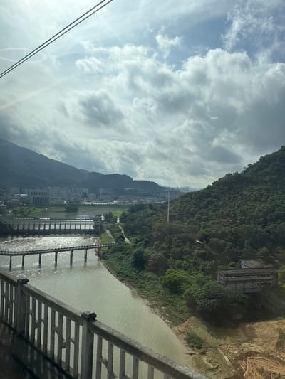 The image captures a view from a moving cable car in Dongguan, China. The scene overlooks a wide river with a prominent concrete dam structure stretching across it. A long, multi-arched bridge crosses the river in the foreground. Beyond the river, a city with numerous buildings is nestled against rolling green hills. Lush vegetation covers the slopes of these hills, with some scattered structures visible amongst the trees. The sky is partly cloudy with dramatic cloud formations, and sunlight filters through, casting rays onto the landscape. The perspective suggests the cable car is traversing a scenic route, likely part of a tourist attraction, offering a panoramic vista of the urban and natural environment of Dongguan.