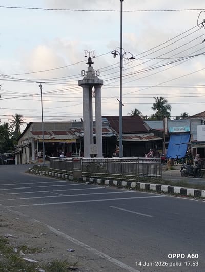 The image depicts a street scene in Uleepulo, Indonesia, featuring a monument with scales of justice on top, flanked by two large pillars. A metal fence separates the monument from the road. Several shops with corrugated metal roofs line the street behind the monument, with awnings providing shade. A few people are visible, including a man in a white shirt and cap standing near the shops, and individuals riding motorcycles. One motorcycle has two passengers, and another is being ridden by a person wearing a headscarf. Several parked motorcycles are also present. The environment appears to be a commercial area with utility poles and overhead wires crisscrossing the sky. The road surface is asphalt with white lane markings. Some litter is visible on the side of the road. The sky is overcast with scattered clouds. The timestamp at the bottom right indicates "OPPO A60 14 Juni 2026 pukul 17.53".