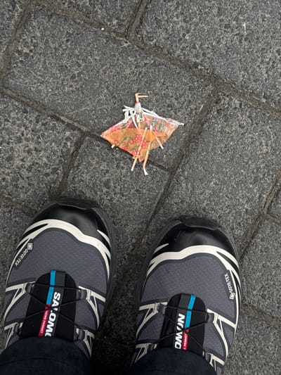 The image presents an overhead view of a pair of Salomon hiking shoes on a stone-paved surface, with a discarded cocktail umbrella on the pavement. The shoes are gray with black and white accents, featuring a quick-lace system and Gore-Tex labeling. The cocktail umbrella is damaged, showing a decorative pattern of flowers. The location can be inferred as Reykjavik, Iceland based on the style of pavement and the common presence of Salomon hiking shoes among tourists.