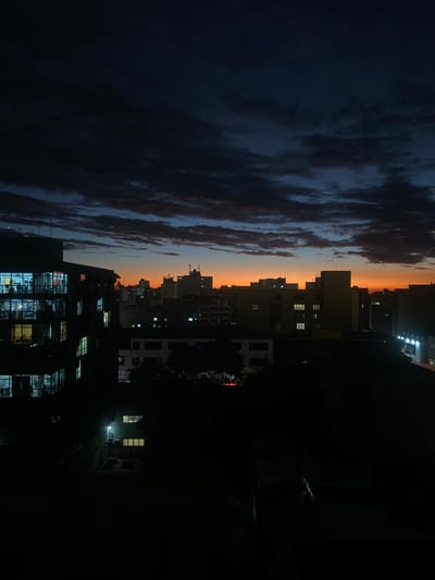 The image presents a panoramic view of a city skyline at twilight, likely dusk, observed from an elevated position. The sky dominates the upper portion of the scene, showcasing a striking gradient of warm orange and yellow hues near the horizon, typical of sunset or sunrise. Above this vibrant band, the sky deepens into shades of dark blue and grey, interspersed with dramatic, elongated, and textured dark clouds that stretch across the frame.
A dense urban landscape unfolds beneath the sky. Numerous buildings form a silhouette against the colorful horizon, their varying heights creating a jagged skyline. In the mid-ground, multi-story buildings are visible, with many windows emitting bright, artificial lights. The immediate foreground features large, dark structures, almost entirely cloaked in shadow, but their numerous windows are brightly illuminated, suggesting active interiors. Some dark, indistinct foliage, possibly trees, can be seen blending into the foreground shadows.
On a rooftop within one of the darker foreground buildings, there are faint points of light and some indistinct shapes that could be equipment or extremely distant human figures, though they are largely obscured by darkness. No specific activity or event is clearly discernible other than the general urban ambiance during the transition from day to night. The scene evokes a quiet, reflective mood. No visible text is present in the image. The location context provided is Caxias Do Sul, Brazil.