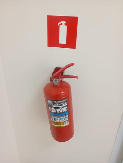 A red fire extinguisher is mounted on a white wall. Above the extinguisher, a red square sign with a white silhouette of a fire extinguisher is displayed. The fire extinguisher itself has a pressure gauge and a red handle. A label with instructions and product information is attached to the body of the extinguisher. The label has text and pictograms illustrating the use of the extinguisher. Visible text on the label includes "OTL-4(S) ABC-01". The setting appears to be indoors, likely in a public space or hallway in Ararat, Armenia. The lighting suggests it is daytime. There are no people present in the image. The overall scene conveys a sense of safety and preparedness.
