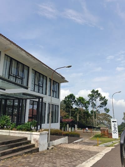 A two-story white building with black-framed windows stands under a partly cloudy sky. A glass awning covers the entrance steps. A rusted street lamp stands near the building. A sign indicating "Raffles" with an arrow pointing left is visible. Trees and a covered structure are in the background, with two people seated. The foreground shows a paved area.