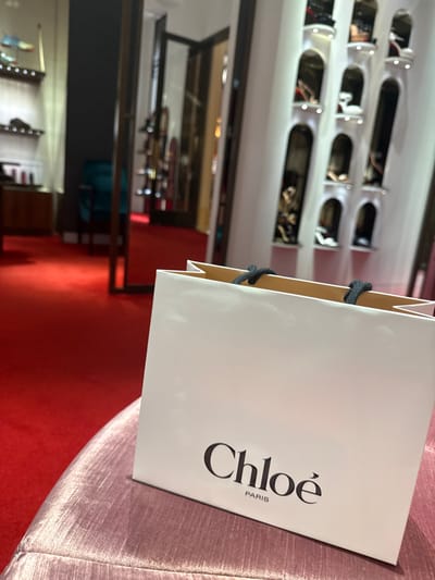 A white Chloé PARIS shopping bag with black fabric handles is positioned on a light pink velvet upholstered surface within a luxury retail boutique, located in Winchester, United States of America. The foreground and midground feature a vibrant red carpeted floor. To the left, a dark-framed doorway leads to an adjacent section of the store where a teal armchair and multi-tiered display shelving with various footwear are partially visible. To the right, a white wall is adorned with multiple vertically aligned, illuminated arched alcoves, each housing a single item of footwear or an accessory for display. No individuals are present in the scene. The environment exhibits high-end retail design with targeted artificial lighting highlighting merchandise.