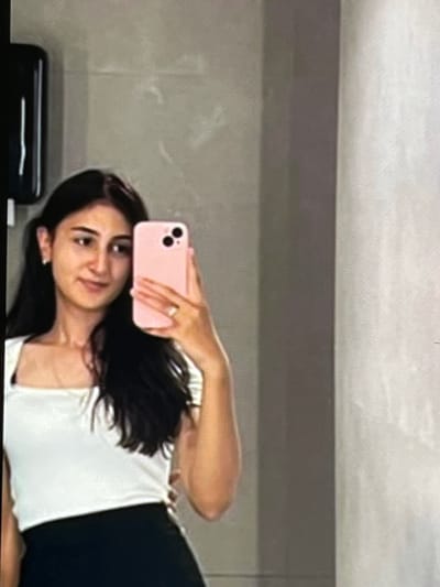 This is a vertically oriented, slightly blurry, close-up selfie taken by a young woman. She is looking towards the camera with a neutral expression. She has dark hair and appears to be wearing a white top and dark bottoms. In her right hand, she is holding a pink smartphone, which is angled to capture the selfie. A ring is visible on her right ring finger.
The background is a plain, neutral-colored wall. A dark object, possibly a piece of furniture or an appliance, is visible on the bottom left of the frame. The overall lighting suggests an indoor setting, possibly a bathroom or dressing room, with no indication of the time of day or weather. No text is visible in the image. The location context is Nijmegen, Netherlands.