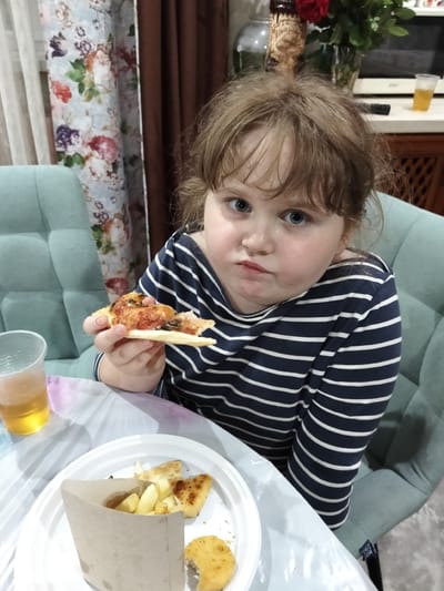 A young child with light brown, tousled hair and blue eyes sits at a table in a room, holding a slice of pizza. The child is wearing a navy and white striped long-sleeved shirt. In front of the child on a white plate are french fries and chicken nuggets. A plastic cup containing a light brown liquid, possibly beer, is to the left of the plate. The table is covered with a white plastic tablecloth. Behind the child, two upholstered chairs are visible, along with a floral curtain and a brown curtain. In the background, a microwave and a vase with red flowers can be seen. The image appears to have been taken indoors, possibly in a home or restaurant setting, in the city of Lobacheve, Ukraine.