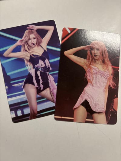 Two K-pop photocards featuring a woman with blonde hair and a pink tint are shown. On the left card, she is wearing a black outfit with pink ruffled trim and a black belt with a heart-shaped buckle. She has her hands near her head and is looking towards the viewer. The background is dark with horizontal blue and white lights. On the right card, she is wearing a sparkling pink corset-like top with a short, light pink skirt over black shorts. She has her hand to her forehead in a salute-like gesture and is looking towards the viewer. The background is dark with red accents. The image is a close-up of these two cards lying on a white surface. There is no visible indication of the city of Velbert, Germany.