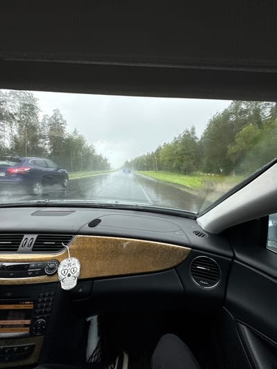 The image is taken from inside a car looking out through the windshield on a rainy day. The windshield is wet with raindrops. Outside, there is a road with cars driving on it. The road is surrounded by trees. The dashboard of the car is visible in the foreground, with a skull-shaped air freshener hanging from the rearview mirror. The location is stated as Гацукоўскі сельскі Савет, Belarus.