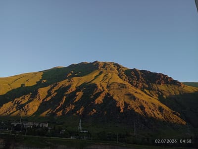 The image depicts a vast mountain range bathed in the golden light of sunrise or sunset. The sun casts long shadows across the slopes, highlighting the rugged terrain and creating a dramatic contrast between light and dark. At the base of the mountain, a small settlement is visible, with buildings and power lines dotting the landscape. The sky is clear and blue, providing a serene backdrop to the majestic scenery. The timestamp "02.07.2026 04:58" suggests that the photo was taken in the early morning of July 2, 2026. The scene is likely located in or near Tyrnyauz, Russia, a city known for its mountainous surroundings and mining history.