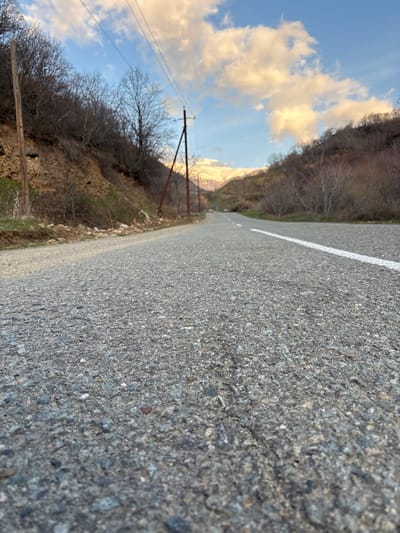 The image depicts a low-angle perspective of an asphalt road in a mountainous, rural area of Gudemnis, Armenia. The road surface, composed of grey aggregate, occupies the foreground and extends into the distance, curving slightly to the right. A solid white line marks the right edge or lane divider of the road.
Flanking the road on both sides are steep hillsides covered with sparse, bare deciduous trees and shrubs, indicative of late autumn or early spring. On the left hillside, closer to the viewer, exposed brown earth and some green ground cover are visible. Two weathered wooden utility poles with overhead lines stand along the left side of the road, receding into the background.
In the far distance, visible at the end of the road's perspective, are snow-capped mountains under a sky featuring blue patches and scattered white clouds tinged with orange and yellow hues, suggesting either sunrise or sunset. No people, vehicles, or animals are present in the scene.