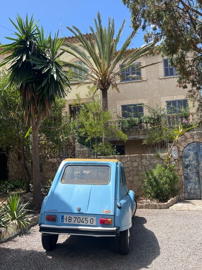 In the city of Pollença, Spain, a classic blue Citroën Dyane 6 is parked on a gravel driveway in front of a two-story tan building with dark blue shutters. A tall palm tree and a yucca plant are visible to the left of the car, with more greenery and trees surrounding the property. The building has a balcony on the second floor, adorned with potted plants. To the right of the building, a rustic stone archway leads to a weathered blue door. The sky is clear and blue. The car's license plate reads "E IB 7045 O".
