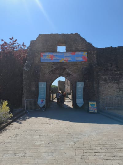 The image depicts the entrance to Ludlow Castle in Ludlow, United Kingdom. The entrance is a large, stone archway, with remnants of the castle walls on either side. A banner is hung across the top of the archway, advertising a series of musical events scheduled for July, featuring artists such as Snow Patrol, Paul Weller, Tom Grennan, and Scissor Sisters.
In front of the archway, people are visible walking through. To the left of the entrance, a lamppost with a blue and white sign for "Ludlow Castle" is present, listing amenities like a cafe, gift shops, and exhibitions. Metal barriers are set up on either side of the pathway leading up to the entrance. A pavement made of stone slabs leads into the castle grounds. The sky is clear and blue, indicating a sunny day. The environment appears to be a historical site with a vibrant event schedule.