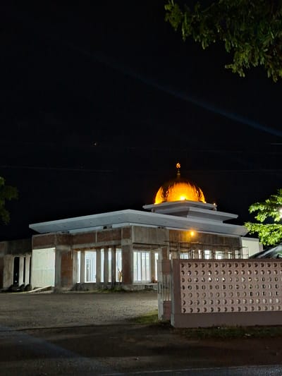 The image captures a nighttime view of a mosque in Reuhattuha, Indonesia, which appears to be under construction or renovation. The central feature is a prominent, brightly illuminated golden dome, crowned with a distinct crescent moon and star finial. Beneath the dome, the building exhibits an unfinished state with exposed concrete beams and pillars, and partial brickwork. The upper roof sections are flat and light-colored, presenting a tiered appearance. The lower section of the building displays multiple vertically oriented white-framed windows, through which interior lighting is visible. Scaffolding-like structures are present on the upper left portion of the building.
The foreground consists of a dark, unpaved ground surface, possibly gravel. To the right, a low, light-colored perimeter wall composed of patterned breeze blocks with circular perforations extends into the frame. Minimal sparse green vegetation is visible at the base of this wall. The sky is dark, indicative of nighttime, and contains faint diagonal light streaks in the upper sections, along with two small, indistinct points of light. Dark green foliage from trees or shrubs partially obscures the view from the upper left and upper right corners of the frame. No individuals or animals are present within the captured scene.