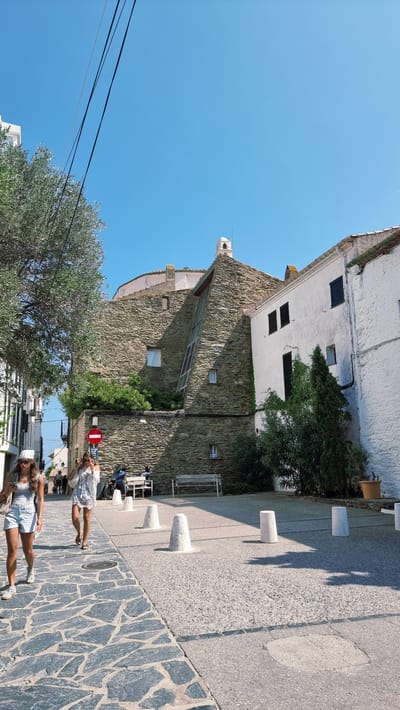 The image depicts a sunny daytime scene in Cadaqués, Spain, with a clear blue sky. Two young women are walking along a cobblestone street lined with white and stone buildings. One woman, in the foreground, wears a white tank top and light blue shorts with white sneakers, accessorized with a white hat. The other woman, further up the street, is in a white, patterned dress and sandals, carrying a straw bag.
The architecture is characteristic of coastal towns, with stone facades and whitewashed walls. Greenery, including an olive tree, adds to the Mediterranean feel. In the background, a motorcycle is parked near a red and white circular "no entry" sign. Several conical bollards are placed along the sidewalk, casting long shadows, indicating the bright sunlight and time of day. The overall atmosphere is relaxed and leisurely, suggesting a vacation or a pleasant stroll.