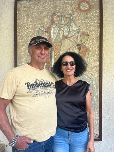 A head-on, daylight photograph captures a smiling man and woman standing in front of a large mosaic artwork, located at Kibbutz Givat Brenner within the Brener Regional Council, Israel. The man, positioned on the left, is fair-skinned with short hair, wearing a beige short-sleeved t-shirt featuring a "Timberland" logo with mountains and trees, blue jeans, and a dark baseball cap with a lighter front detail. He has a multi-strand silver-toned beaded bracelet on his left wrist, with his left hand partially in his pocket. The woman, on the right, has dark, curly hair and wears dark round sunglasses, a black V-neck sleeveless top, and light blue high-waisted jeans with a visible four-button fly. She wears dangling teardrop-shaped earrings. Both individuals smile directly at the camera; the woman's right arm is positioned behind the man's lower back. Behind them, the mosaic, set within a dark brown wooden frame, depicts abstract, angular figures in tones of brown, orange, grey, and white against a lighter, speckled background, with a circular sun-like motif in the upper right. A textured, light-colored wall is visible to the left of the man and alongside the mosaic.
