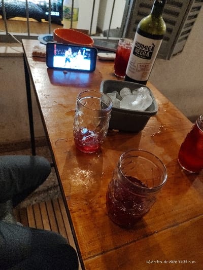 The image depicts a late-night scene on a wooden table in Bernal Este, Argentina, as indicated by the timestamp "28 de feb. de 202x 11:3x p.m.". A person, visible only by their legs in dark pants in the lower left, is seated at the table. Central to the table is a horizontally oriented smartphone displaying a video of what appears to be a basketball game. To the left of the phone, an empty red-orange bowl rests on the table. To the right, a bottle of "HORMIGA NEGRA MALBEC" wine stands next to a grey container filled with ice cubes. Four glasses are present: one mostly empty with dark red residue near the phone, one textured glass partially filled with dark red liquid in the foreground, another partially visible behind the wine bottle also containing dark red liquid, and a fourth partially visible on the far right, also containing dark red liquid. The wooden table surface shows multiple liquid spills and condensation rings. In the blurred background, through a barred opening, artificial lighting is visible.
