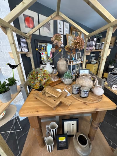 This image depicts a detailed interior view of a retail establishment, identified as being located in Hornsea, United Kingdom, specifically a home goods or gift shop. The scene is dominated by a light-colored wooden structure resembling a house frame, under which a substantial light-brown wooden table is positioned centrally.
On the table, a curated display of merchandise is arranged. Objects include: several rectangular wooden serving boards, some with handles and raised feet; two large, textured, grey-brown ceramic pitchers or vases; two cylindrical grey scented candles in jars with metallic lids, placed on a round wooden serving board; a small, pale pink ceramic vase; a clear glass bowl featuring a multi-colored, possibly floral or leaf, design; a small green succulent plant in a grey pot; two empty stemmed glass coupes; and a diffuser bottle with reeds. A small black bird figurine on a green stake is positioned near the glass bowl.
The background features a dark grey-blue wall adorned with various framed artworks, including minimalist abstract prints, botanical illustrations, and a framed "Bauhaus" poster. A large, dark-framed mirror on the back wall reflects a partial view of another section of the store, showing indistinct shapes and a white chair. To the left, a white brick wall segment is visible, with wooden shelves holding two small potted green plants in grey metal buckets and another black bird figurine on a green stake. Above, bags of merchandise are on a shelf. To the right, additional shelving displays various ceramic items, and a yellow chest of drawers is partially visible below.
Beneath the central wooden table, two black and white striped ceramic pitchers, two small framed artworks (one featuring the word "Bauhaus" and the other an abstract yellow flower), and a large textured grey ceramic vase are displayed directly on the dark tiled floor. The floor consists of large dark grey tiles interspersed with lighter grey geometric patterns. No individuals are visible in the scene. The lighting is bright and even, indicating an illuminated interior space.